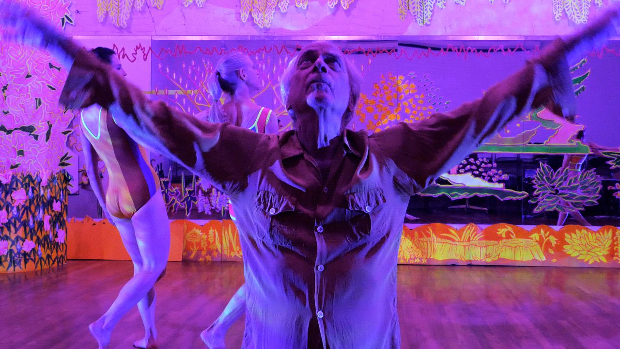 White-haired white man with arms uplifted and face to ceiling bathed in purple light in front of an exuberantly painted mirror in the background